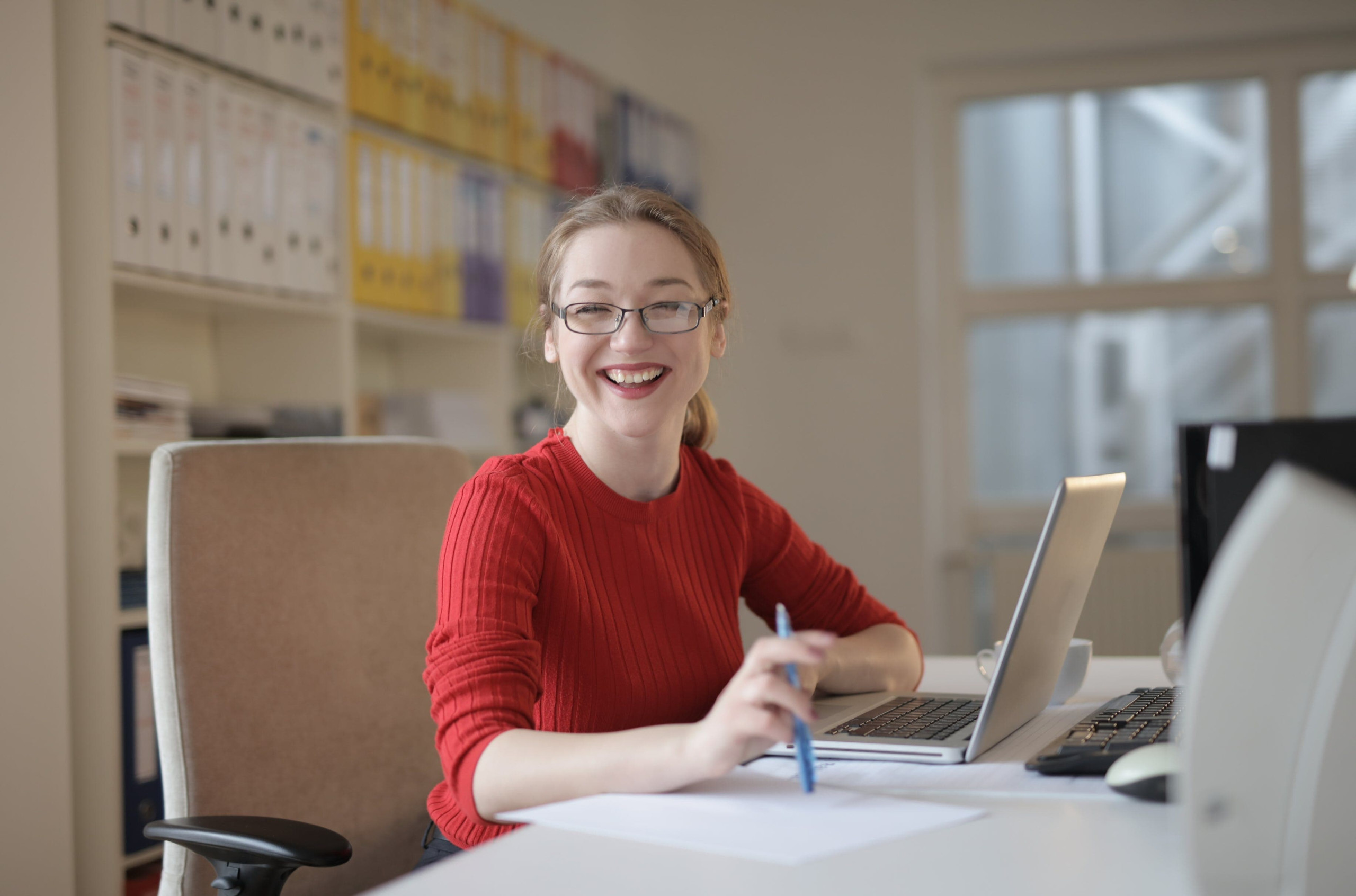 A women with a red top sitting at a desk with a laptop and smiling at the camera